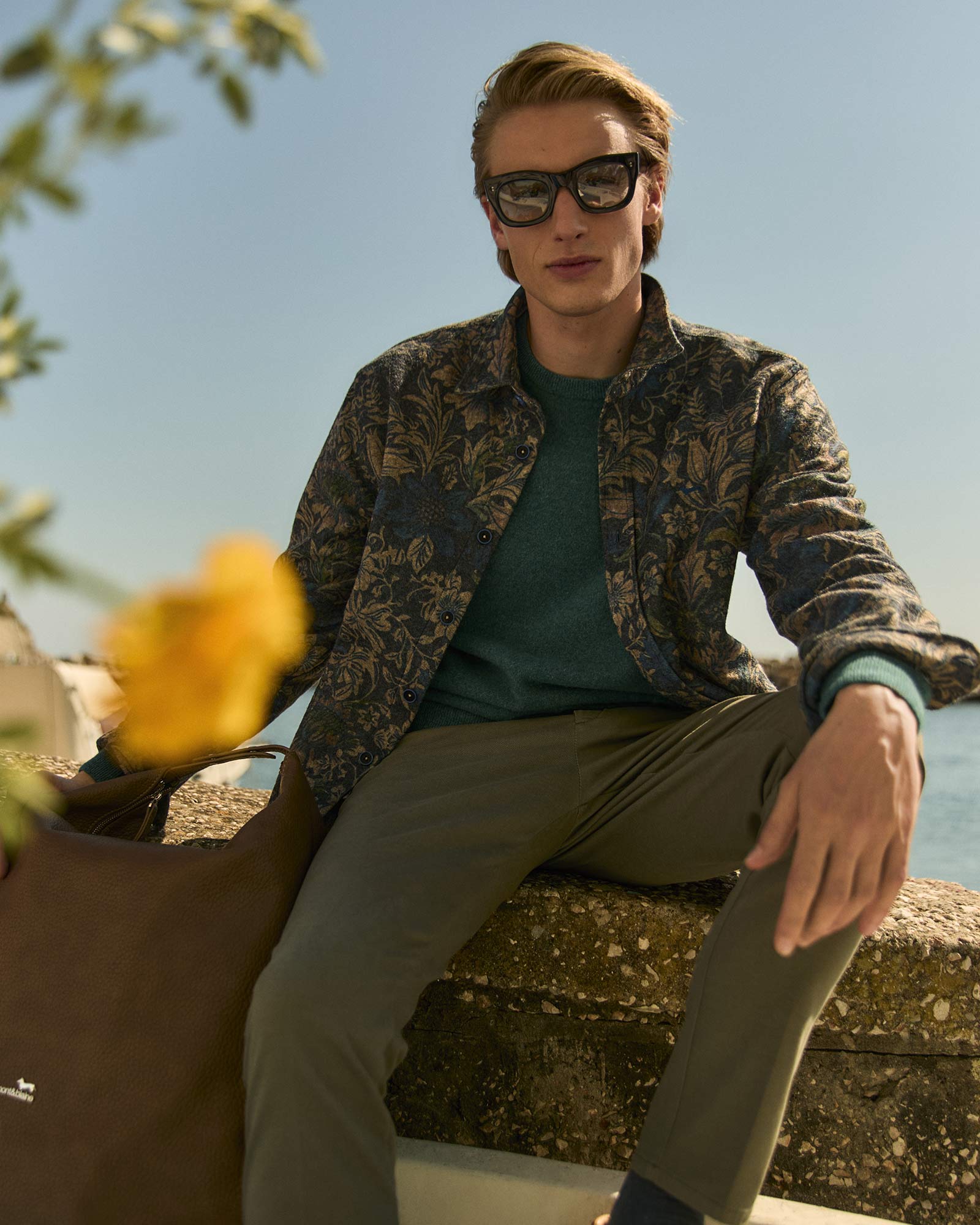 Uomo con camicia a fantasia floreale e occhiali da sole, seduto all’aperto in riva al mare.